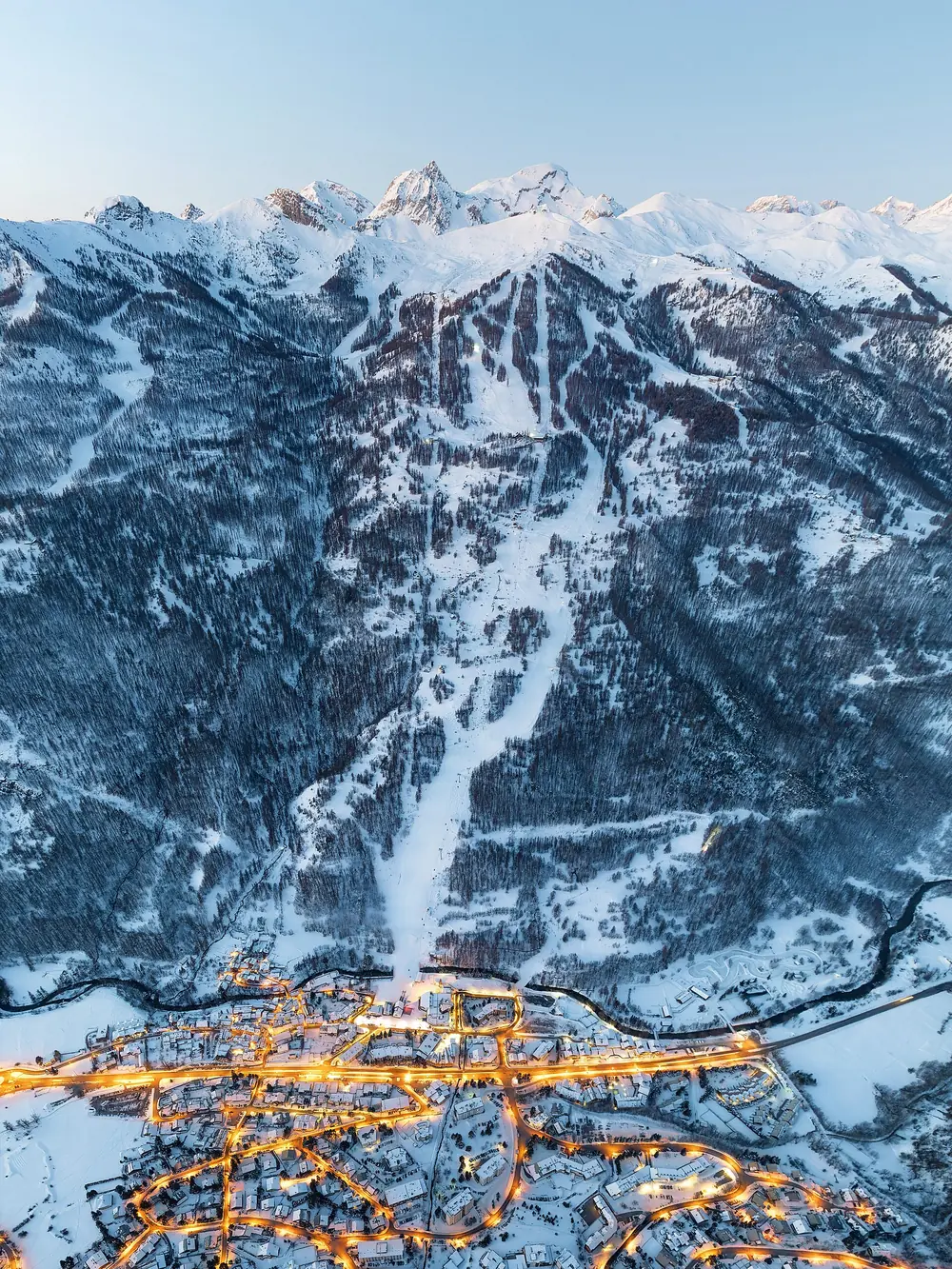 Chantemerle à l'heure bleue en décembre, vue aérienne : village éclairé, pistes enneigées en forêt et sommets au fond